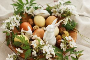 Modern easter eggs with spring flowers and bunny in wooden bowl on rustic linen cloth. Happy Easter! Natural dyed eggs on grey textile with blooming flowers anemone and hare. Aesthetic