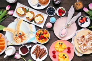 Fun Easter breakfast or brunch table scene. Top down view on a dark wood background. Bunny pancake, egg nests, chick fruit and assorted spring food items.