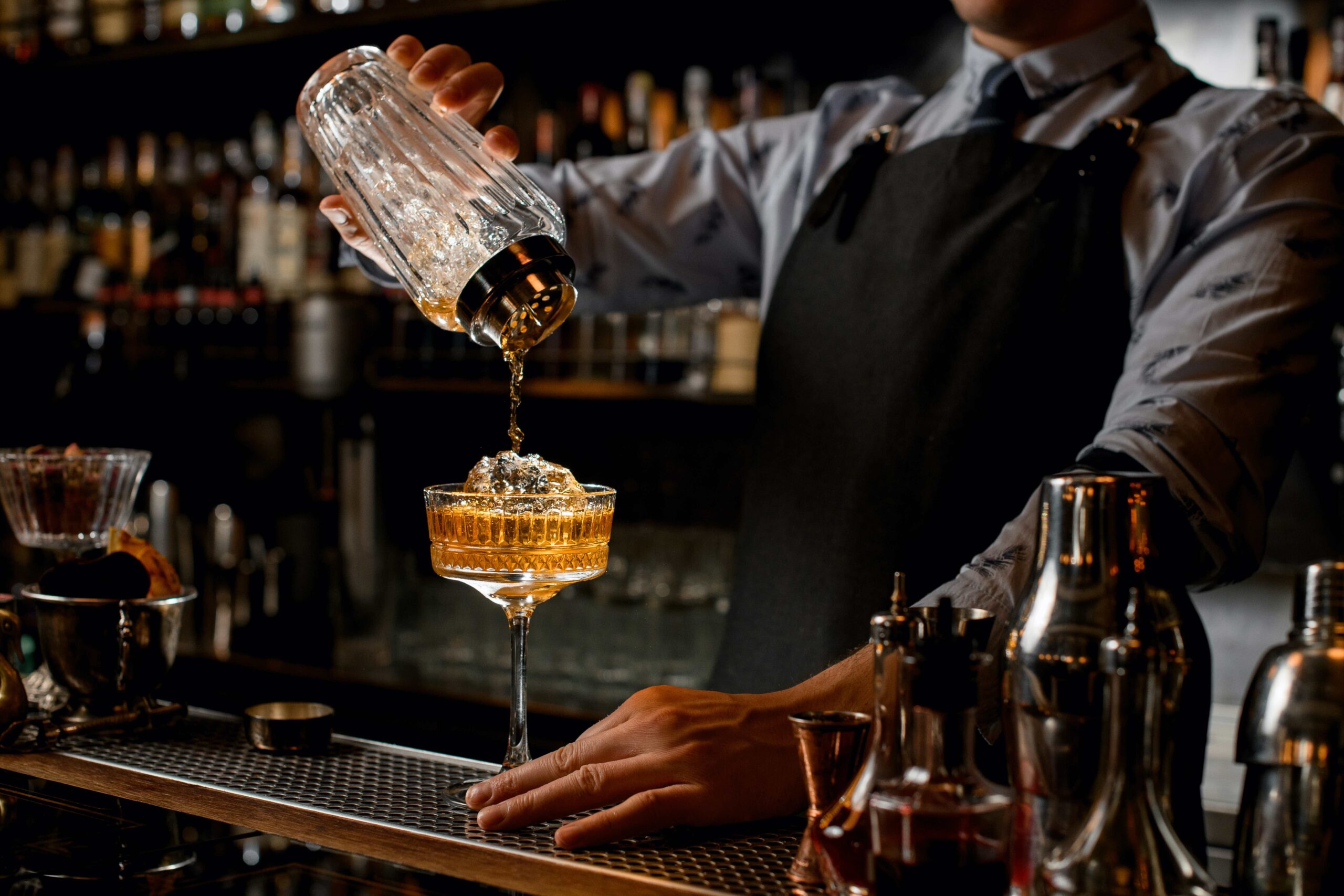 Professional bartender in black apron pours brown drink from shaker into glass