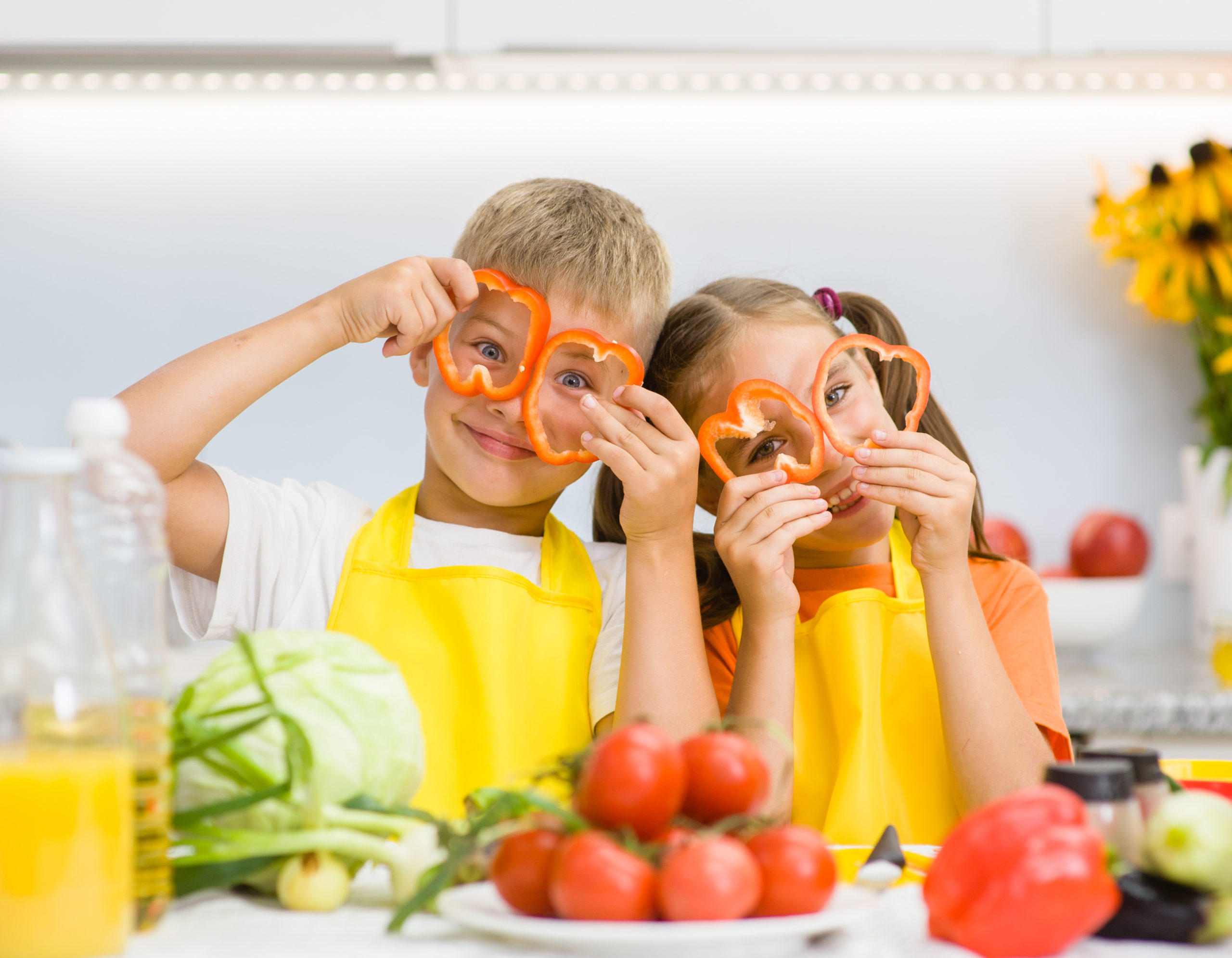 Two children with vegetables and peppers holding the peppers over their eyes like glasses