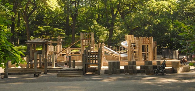 A wooden playground surrounded by large green trees on a sunny day in Central Park.