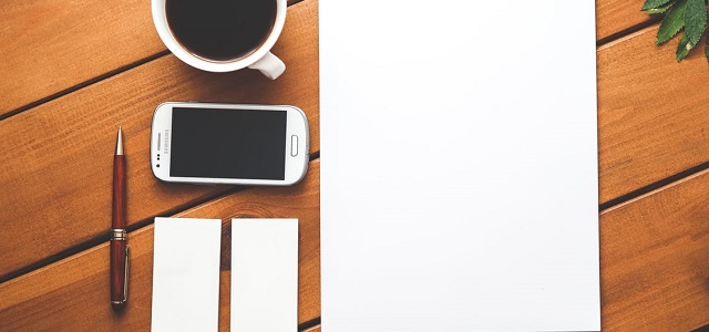 A white piece of paper, red pen, coffee, and smartphone sit on a brown wooden table.