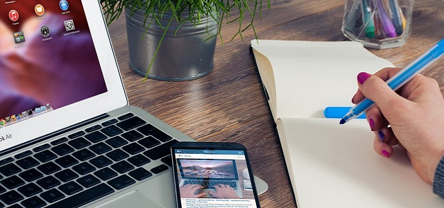 Photo Credit: A woman's hand holding a blue marker with a white pad of paper and laptop on a wooden table.