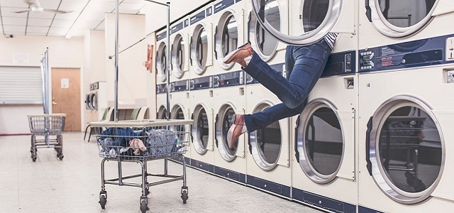 A man hanging from a washing machine searching for missing laundry in a laundromat.