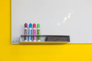 A whiteboard on a yellow wall with colorful markers