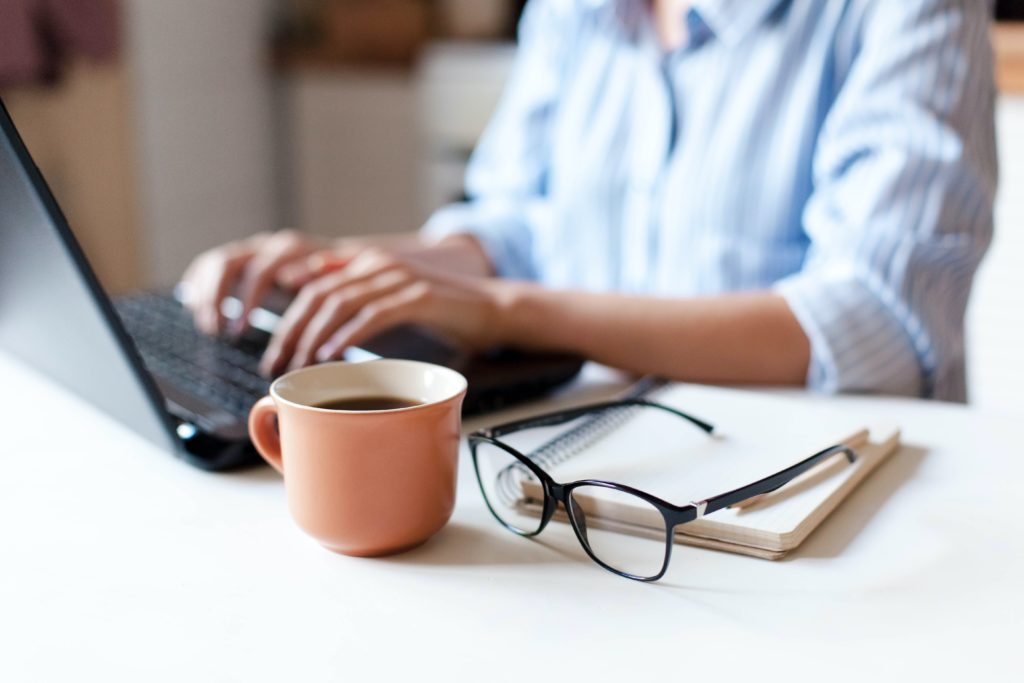A women working from home at her desk with a laptop, notebook, glasses and coffee mug