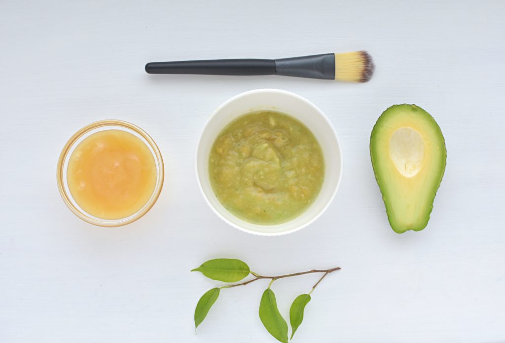 Avocado and honey mixed in a bowl with a makeup brush and leaf on a white backgronud