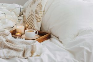 Wooden tray of coffee and candles on bed. White bedding sheets with striped blanket and pillow. Breakfast in bed.