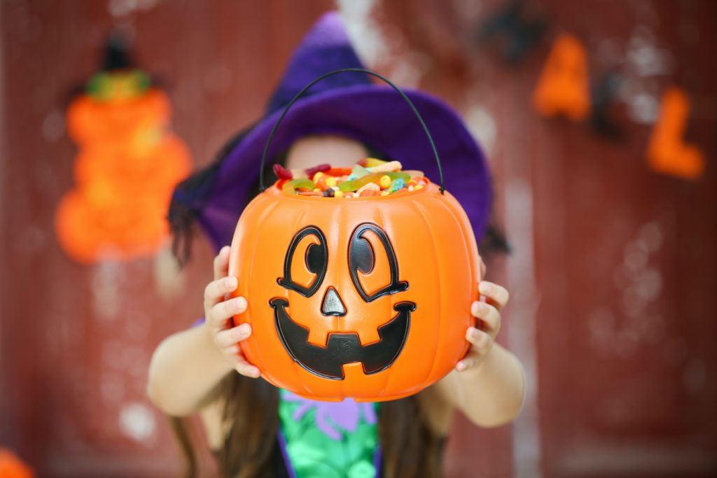A child with a purple witch hat holding a trick or treating pumpkin out over her face filled with candy.