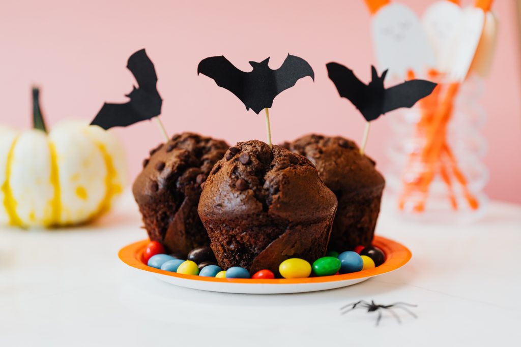 Halloween chocolate muffins with bats stuck with toothpicks, a small pumpkin and ghost straws in the background.