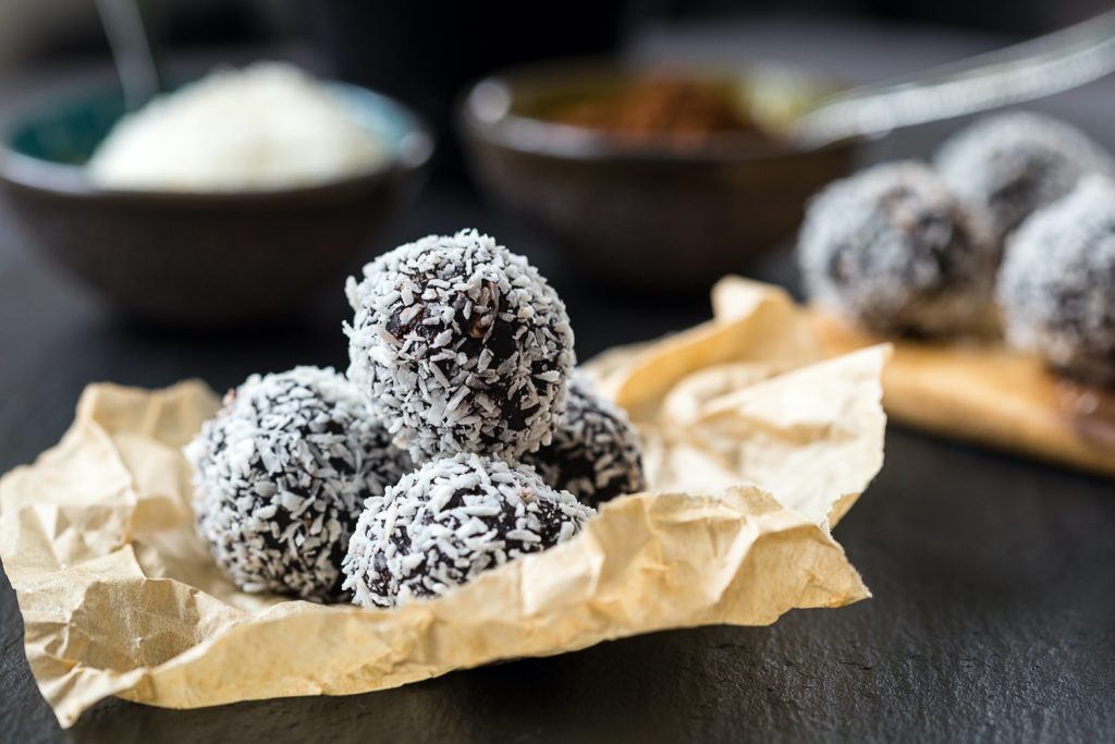 Coconut truffles on parchment paper, in the background two bowls one with coconut flakes the other with coco powder