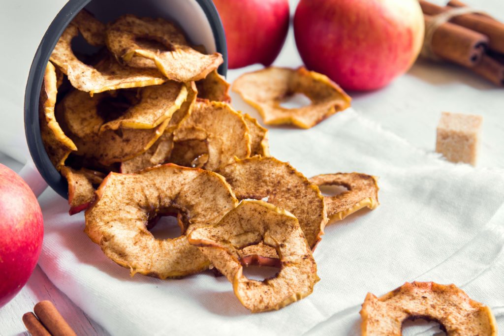 A mug with cinnamon apple chips coming out on a table with apples, brown sugar cubes and cinnamon sticks