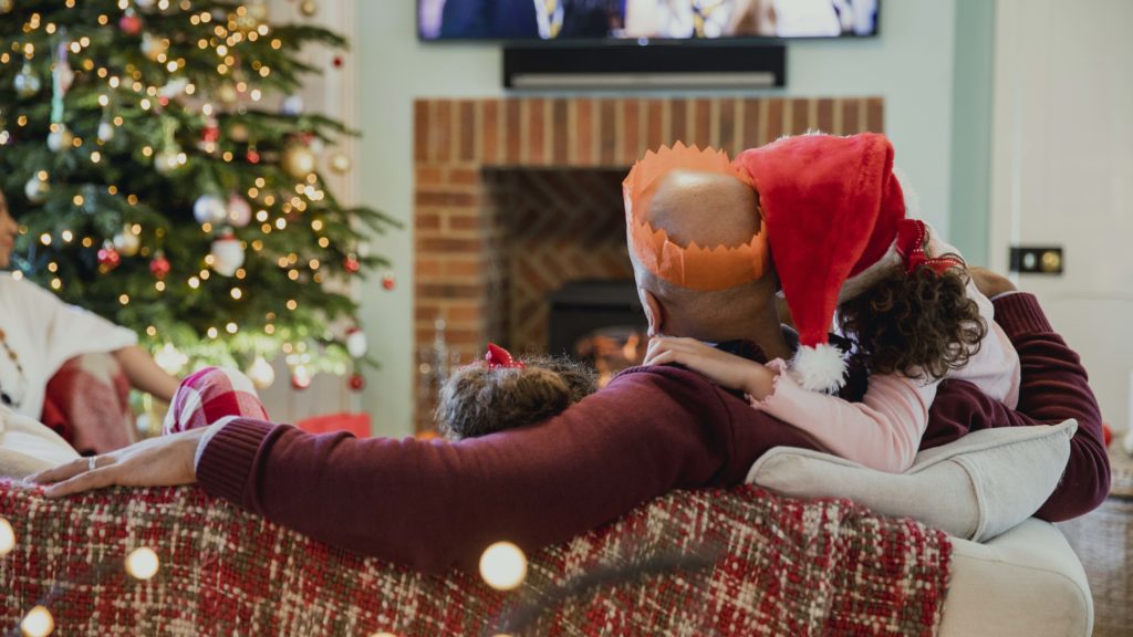 A father curled up on the couch watching a movie with his two daughters one in a Christmas hat