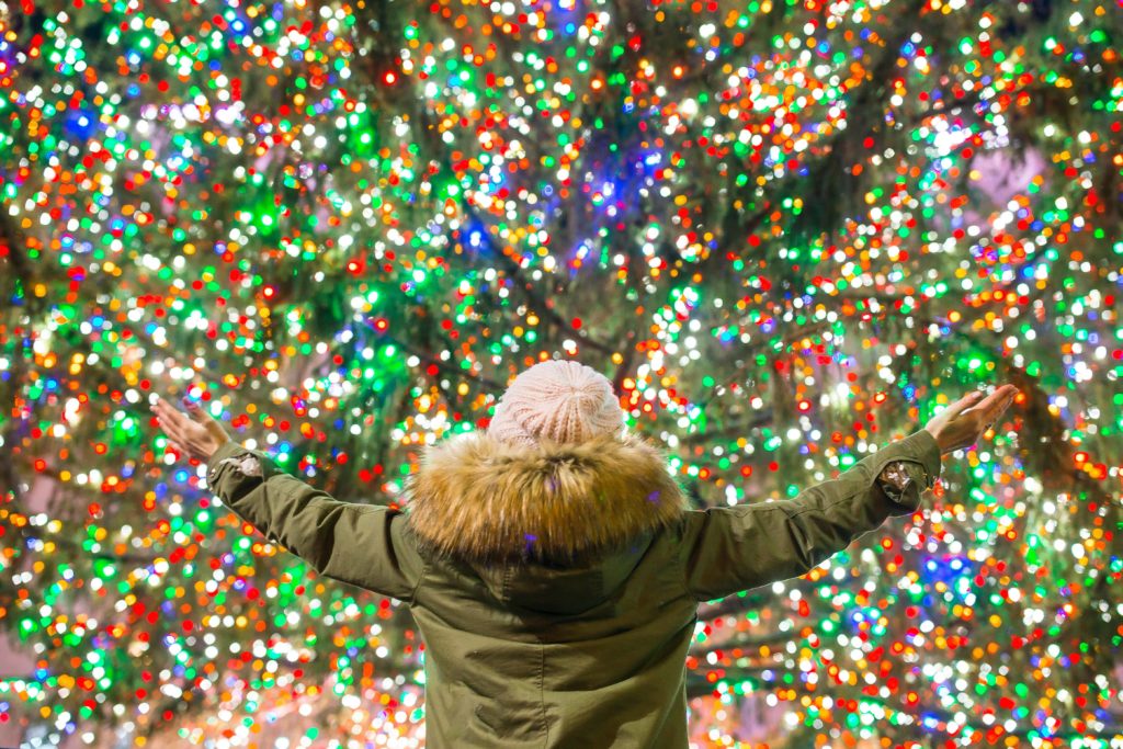 A close up shot of a woman standing in front of the lit up Rockefeller Center Tree with her hands raised