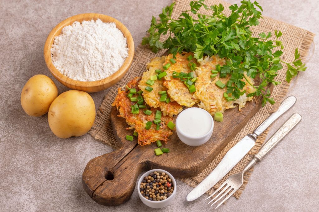 potato latkas on a cutting board with a side of sour cream surrounded by two potatoes, a bowl of flour, parsley and silverware