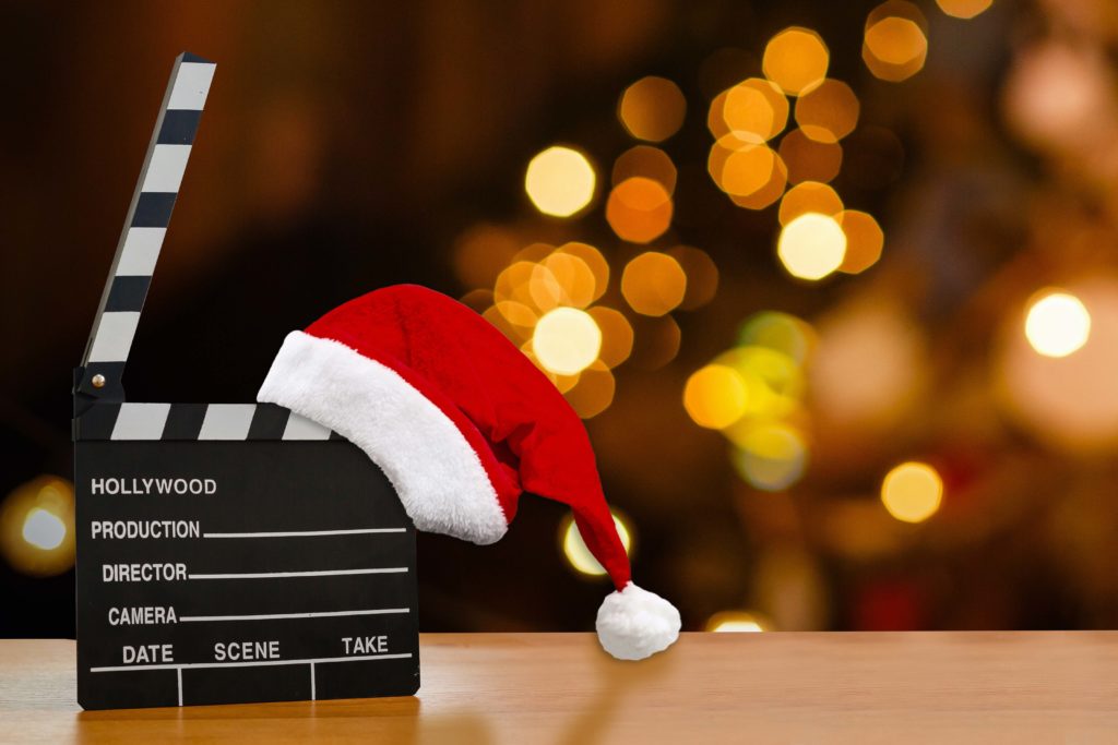 A movie clap board with a Santa hat on it sitting on a wood table with twinkling lights behind it