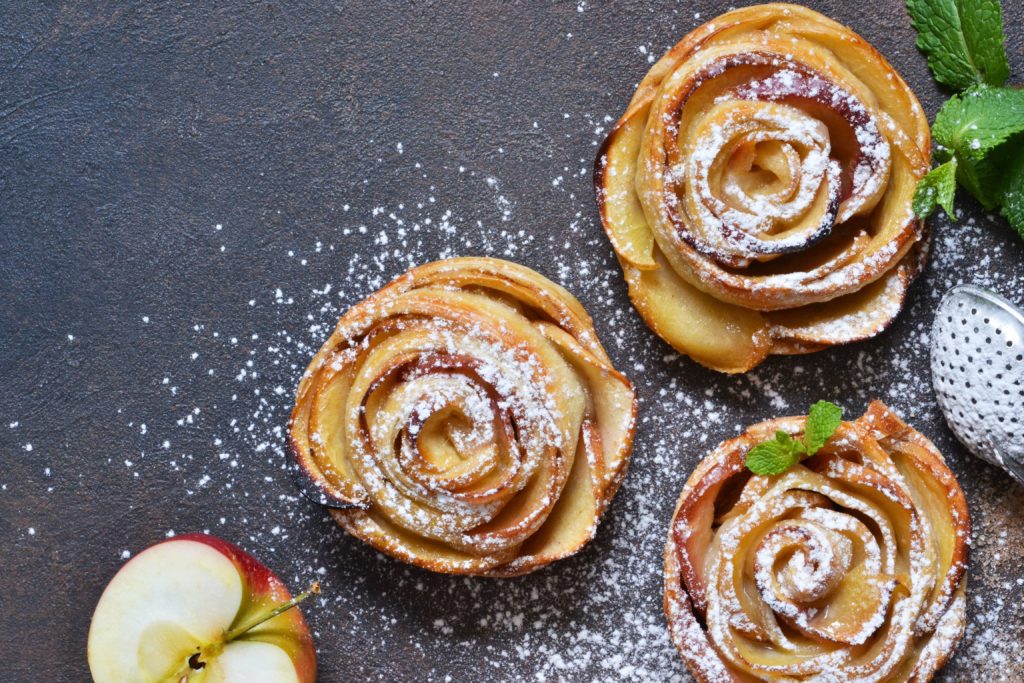 3 apple flower tarts on a black background with half an apple and sprinkled with powdered sugar
