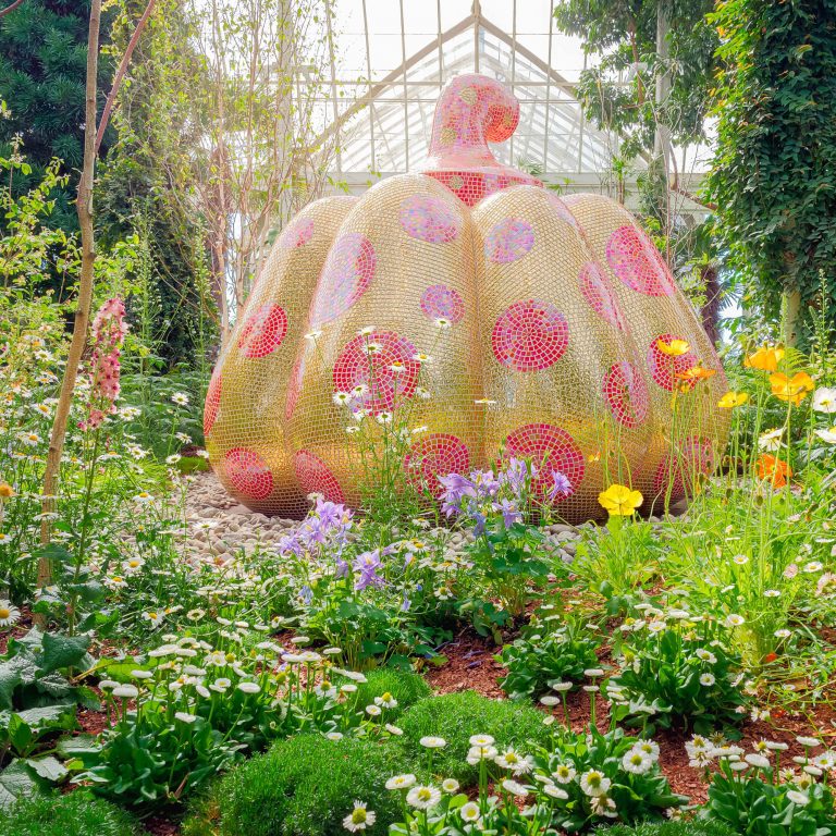 A bedazzled yellow and pink Kusama pumpkin sculpture amongst beautiful flowers