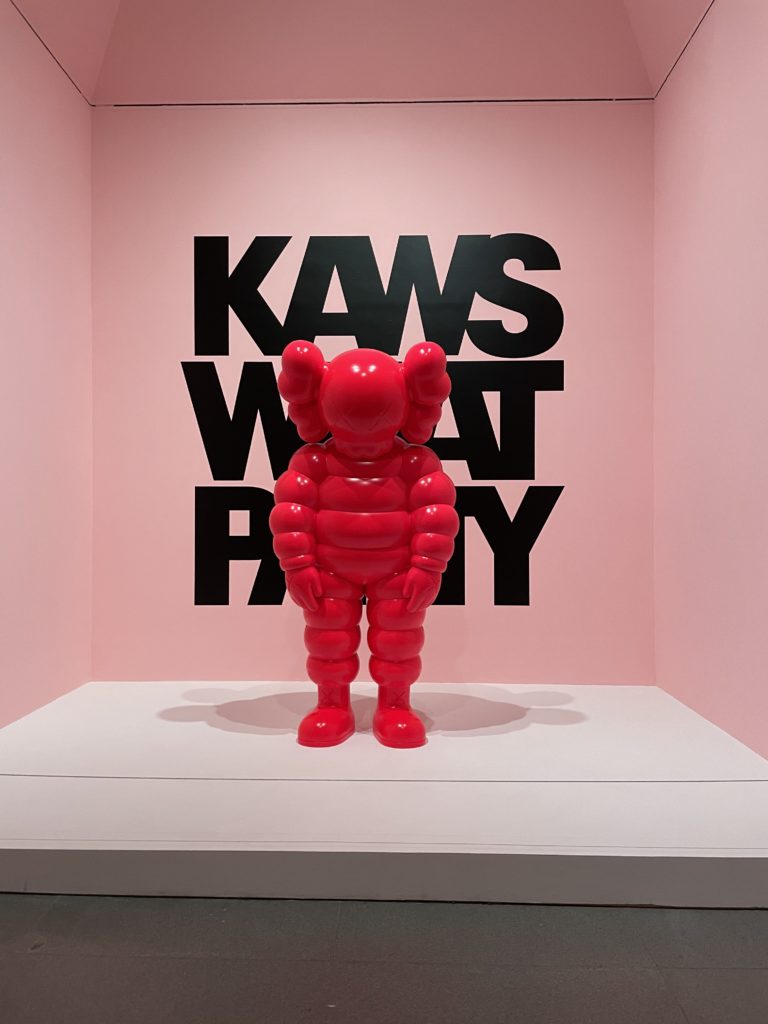 Large pink KAWS in front of a pink KAWS: WHAT PARTY background