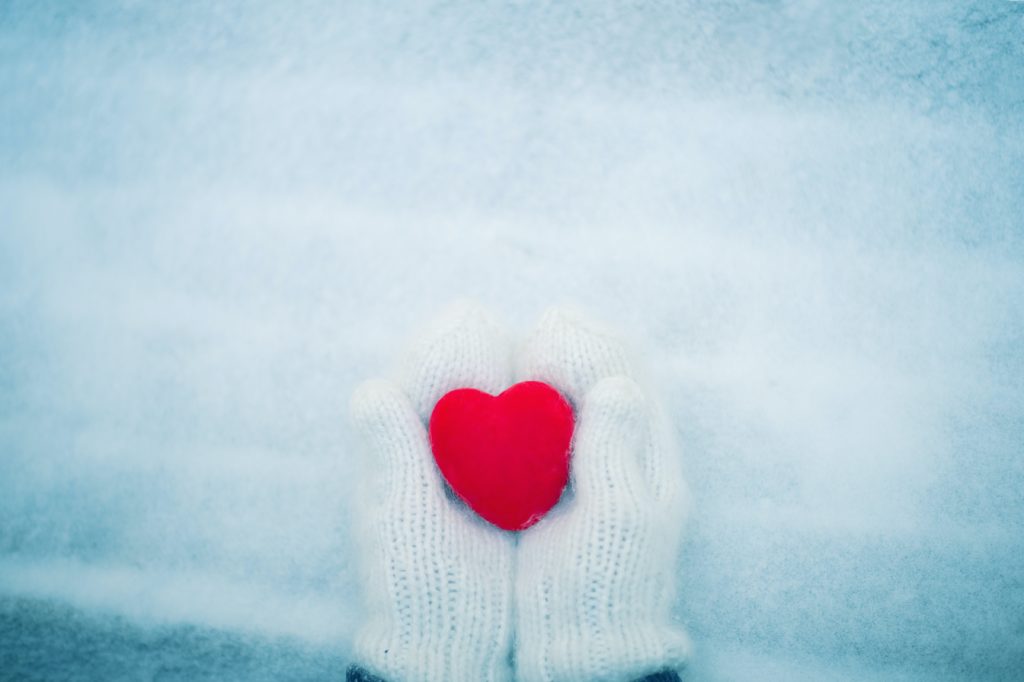 Red heart in woman's hands wearing white woolen mittens. Top view snow background
