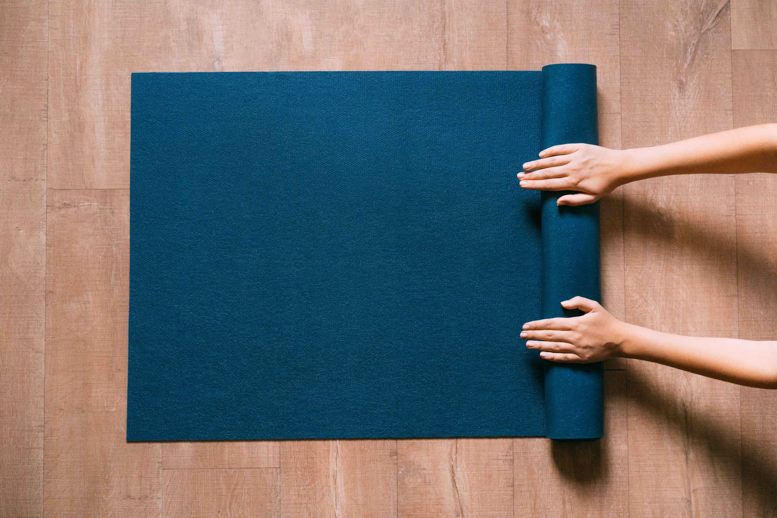 woman rolling up a blue yoga mat on a wood floor