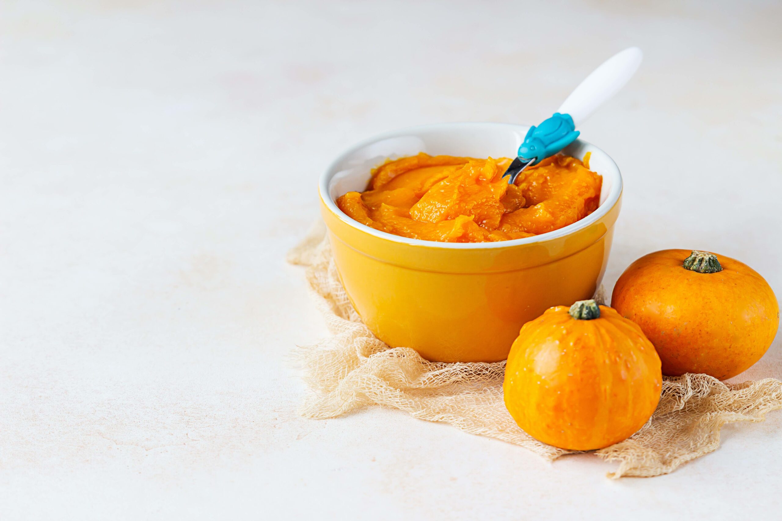 Homemade pumpkin puree in bowl with baby spoon and fresh pumpkins on light background. The concept of baby food.