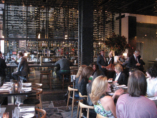Interior view of Colicchio and Sons NYC restaurant in the meat packing plant filled with glass tables, wood chairs, a large bar adorned with a wine rack and diners