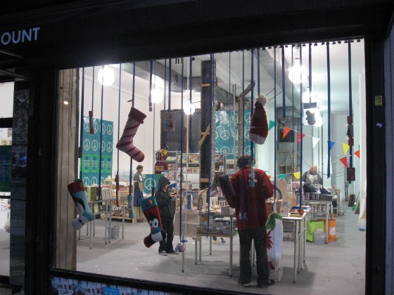 view from the street of holiday shopping pop up the Blue Ribbon General Store with stocking and hats tied to streamers dangling in front of the window and shoppers in the background