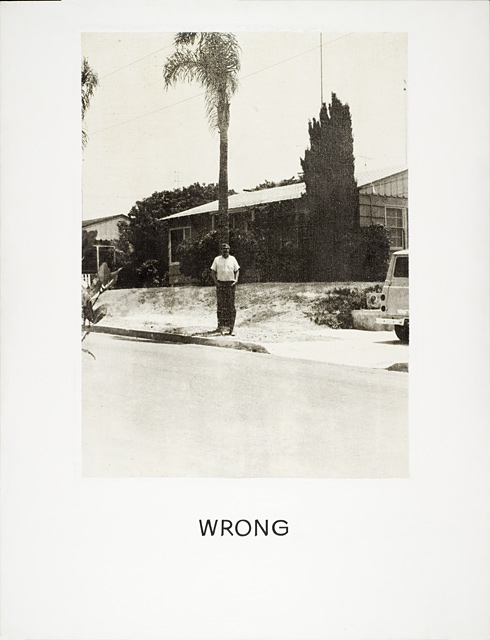 John Baldessari photograph of a man in a white t-shirt with dark pants in fron of a house set behind a palm tree so the pants fade into the tree