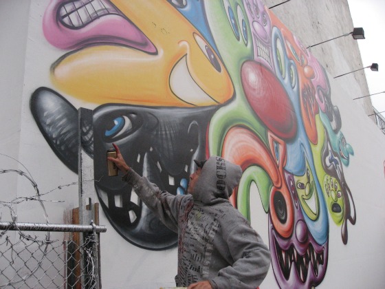 Artist Kenny Scharf working on his Houston Street mural can of spray paint in hand in a grey hoodie painting cartoon faces