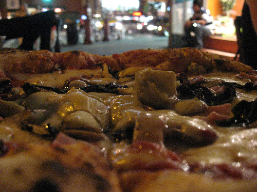 Olio Pizza NYC close-up of a pizza pie with cheese, proscuitto, fresh basil, and tomato sauce on a plate with the streets of Manhattan in the background