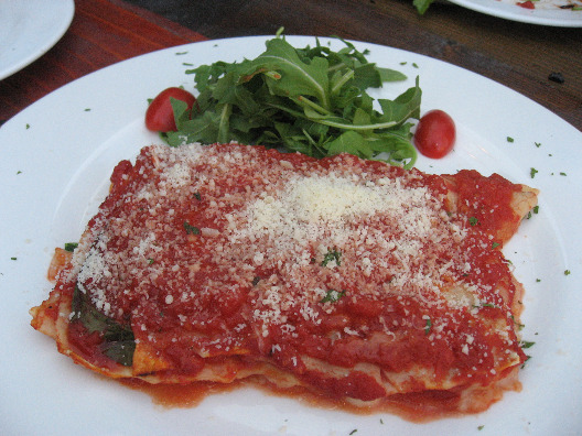 Olio Pizza lasagna on a white plate drenched in fresh tomato sauce, sprinkled with cheese and a small arugala and tomato salad on the side