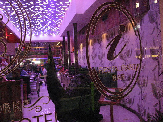 View from outside into Restaurant i with logo on the window and purple interior, colorful ceiling, flowered wallpaper, and long line of tables and chairs